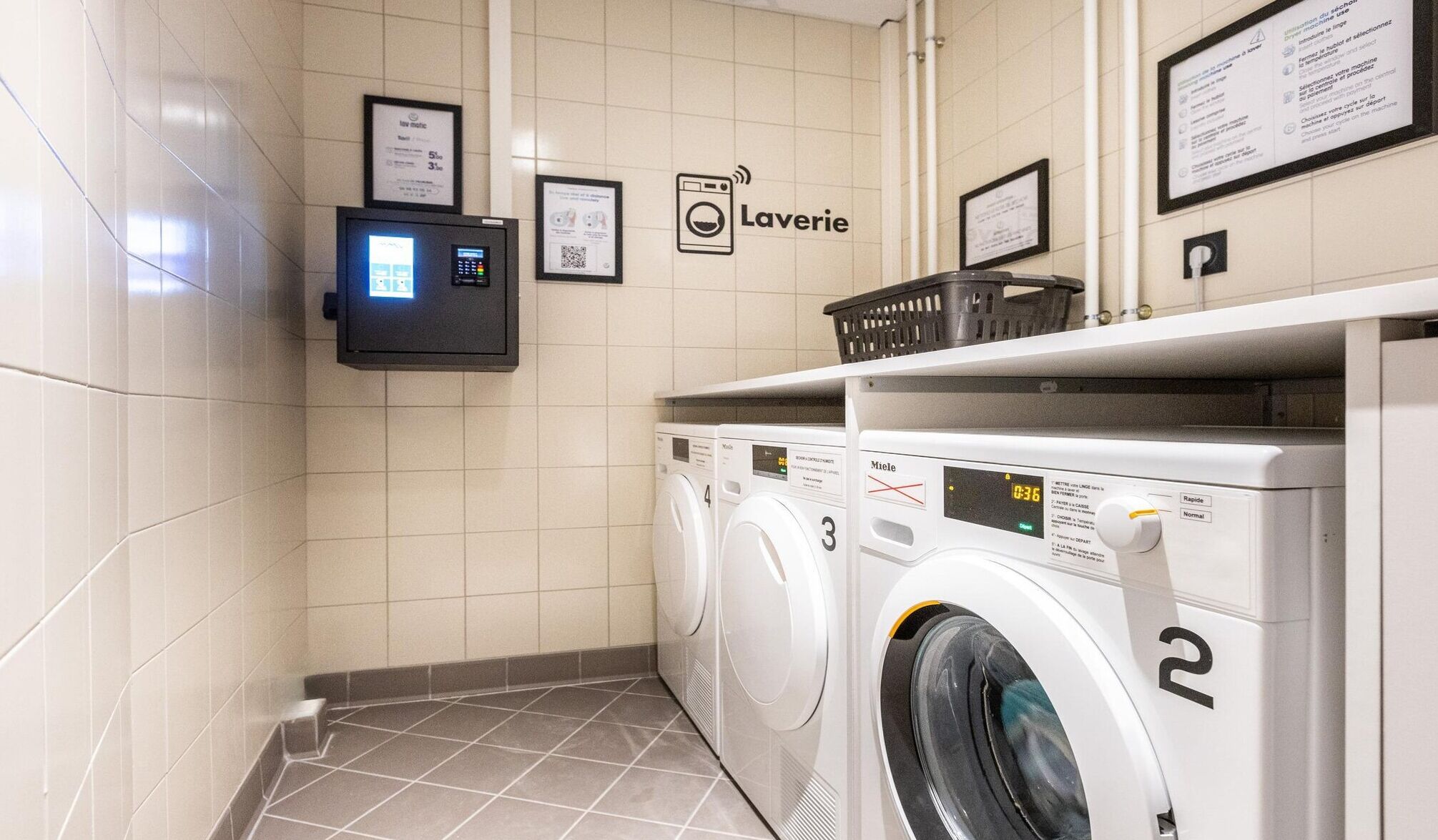 Una lavanderia self-service con tre lavatrici e asciugatrici, un pannello di controllo digitale e cartelli informativi alle pareti.