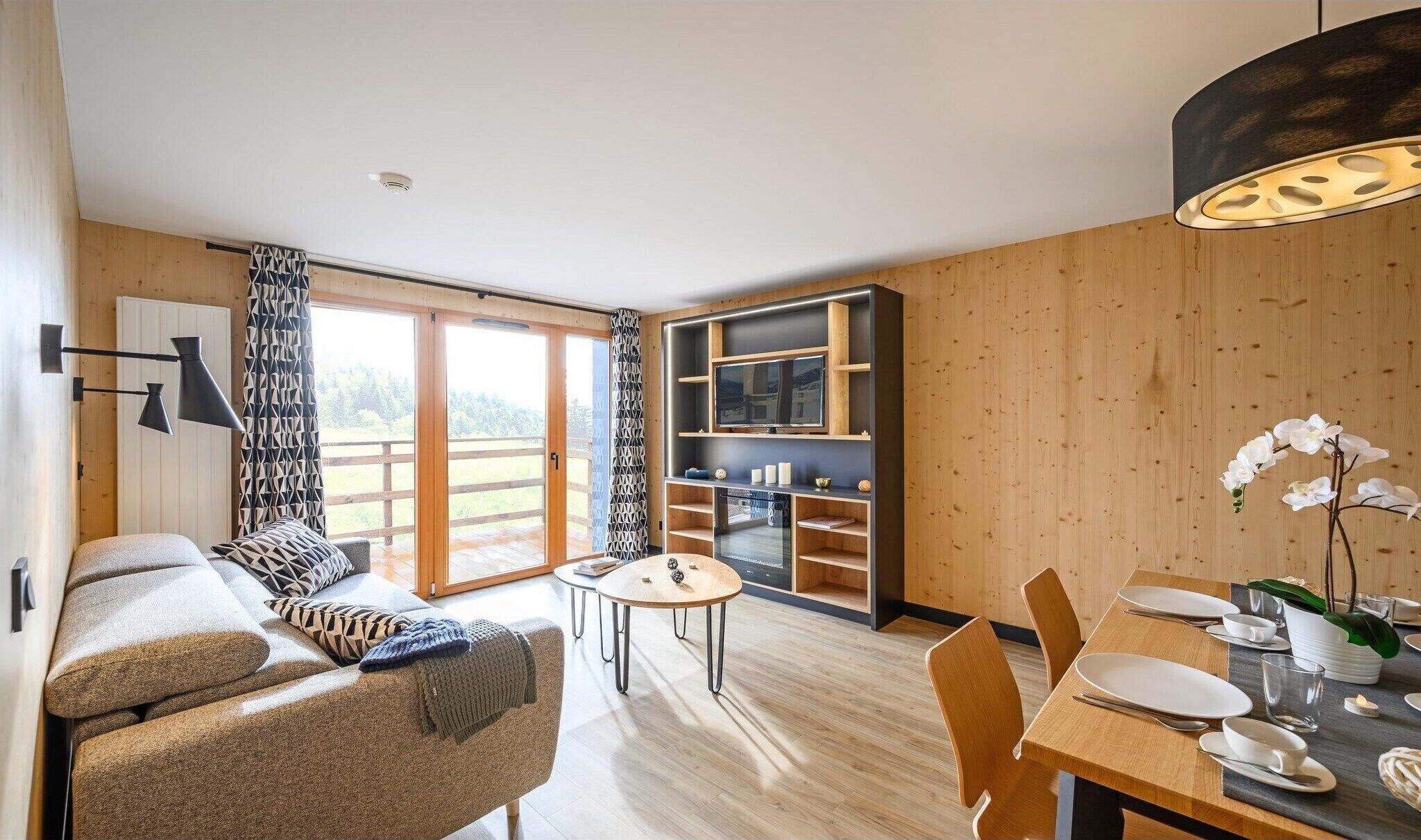 A modern living room with a sofa, a dining table set for four, and a kitchen area with wooden cabinets and a built-in oven.