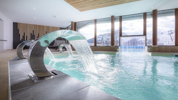 A modern indoor swimming pool with a unique water feature, large windows, and a view of snow-capped mountains.