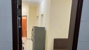 Ein Zimmer mit Fliesenboden, einem grauen Kühlschrank, einem braunen Sofa und einer Tür zu einem weiteren Raum.