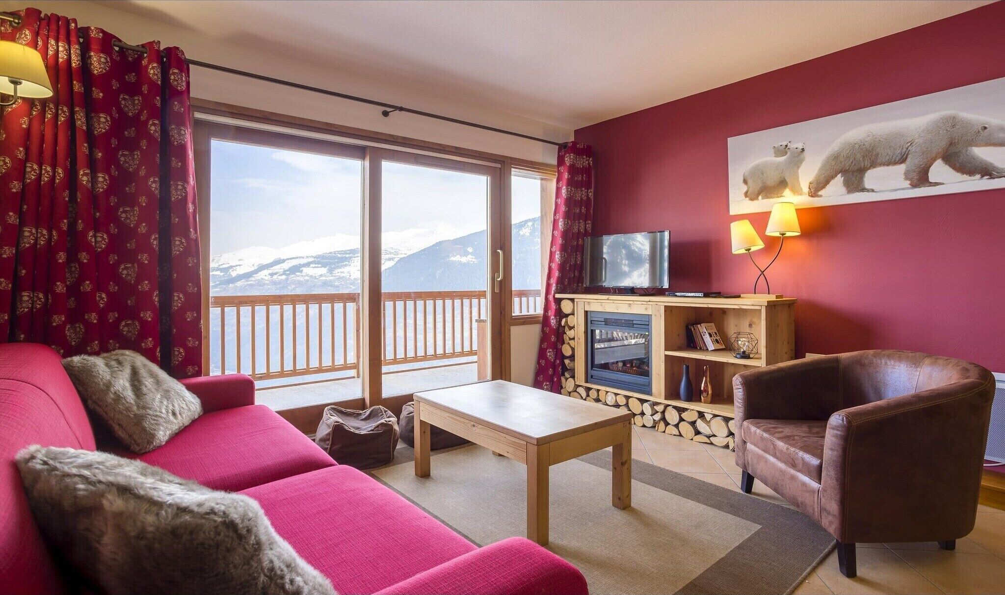 Een gezellige woonkamer met een rode bank, een bruine fauteuil en een houten salontafel. Een televisie staat op een houten dressoir en er is een groot raam met uitzicht op besneeuwde bergen.