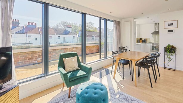 Ein modernes Wohnzimmer mit großem Fenster, einem grünen Sessel, einem gepolsterten Hocker, einem Holzespeisetisch und einem Fernseher auf einem Holzständer.