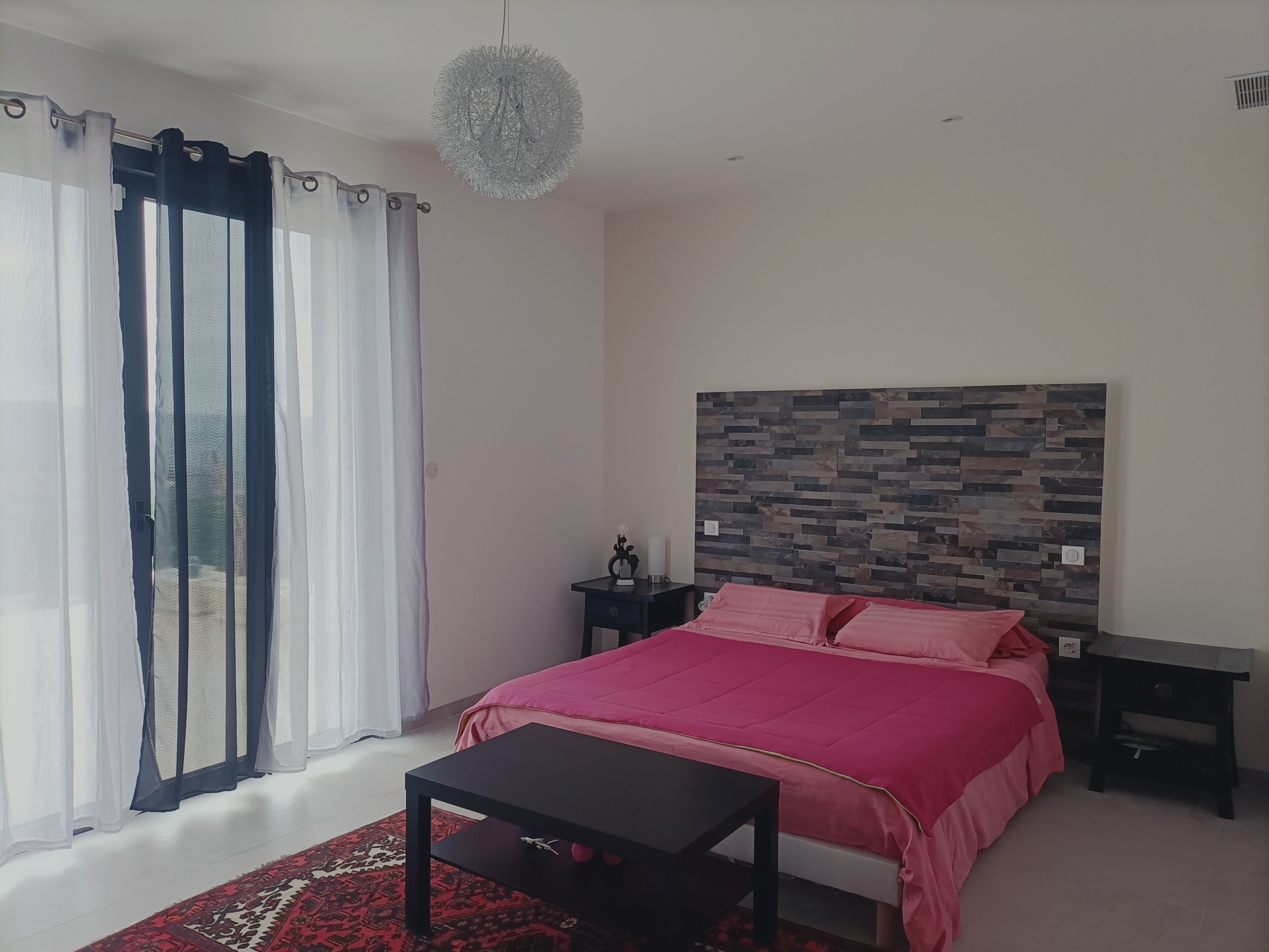 Un dormitorio con cama, mesita de noche, mesa, candelabro y vistas a través de una ventana con cortinas.