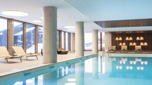 Een modern binnenzwembad met helderblauw water, omringd door ligbedden en lounge fauteuils, grote ramen en een houten plafond.