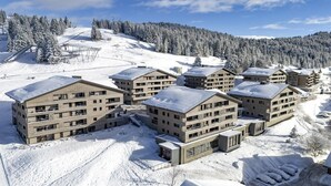 Een met sneeuw bedekte skigebied met moderne, meer verdiepingen tellende gebouwen, skiliften en een bebost berglandschap op de achtergrond.