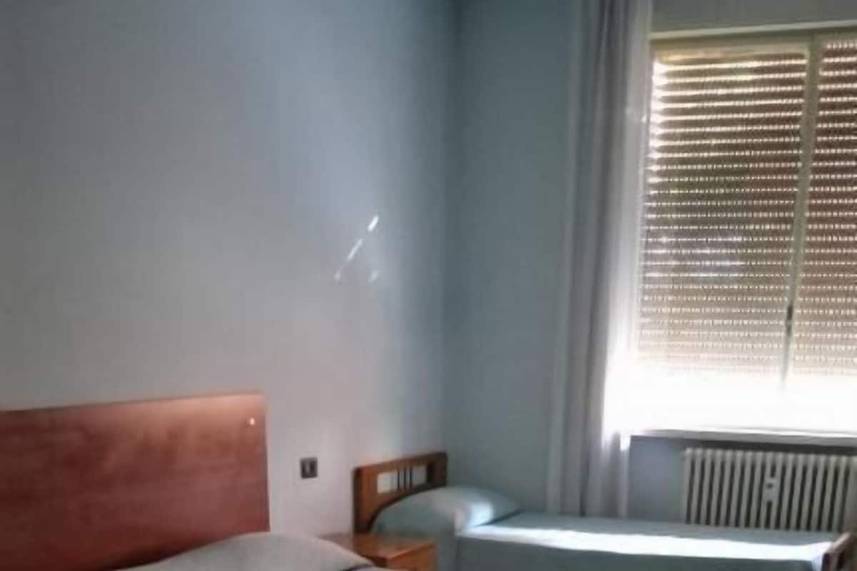 Quarto com três camas alinhadas, radiador na parede e janela com persianas.
