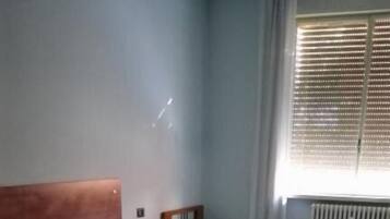 Una habitación con tres camas dispuestas en fila, un radiador en la pared y una ventana con persianas.