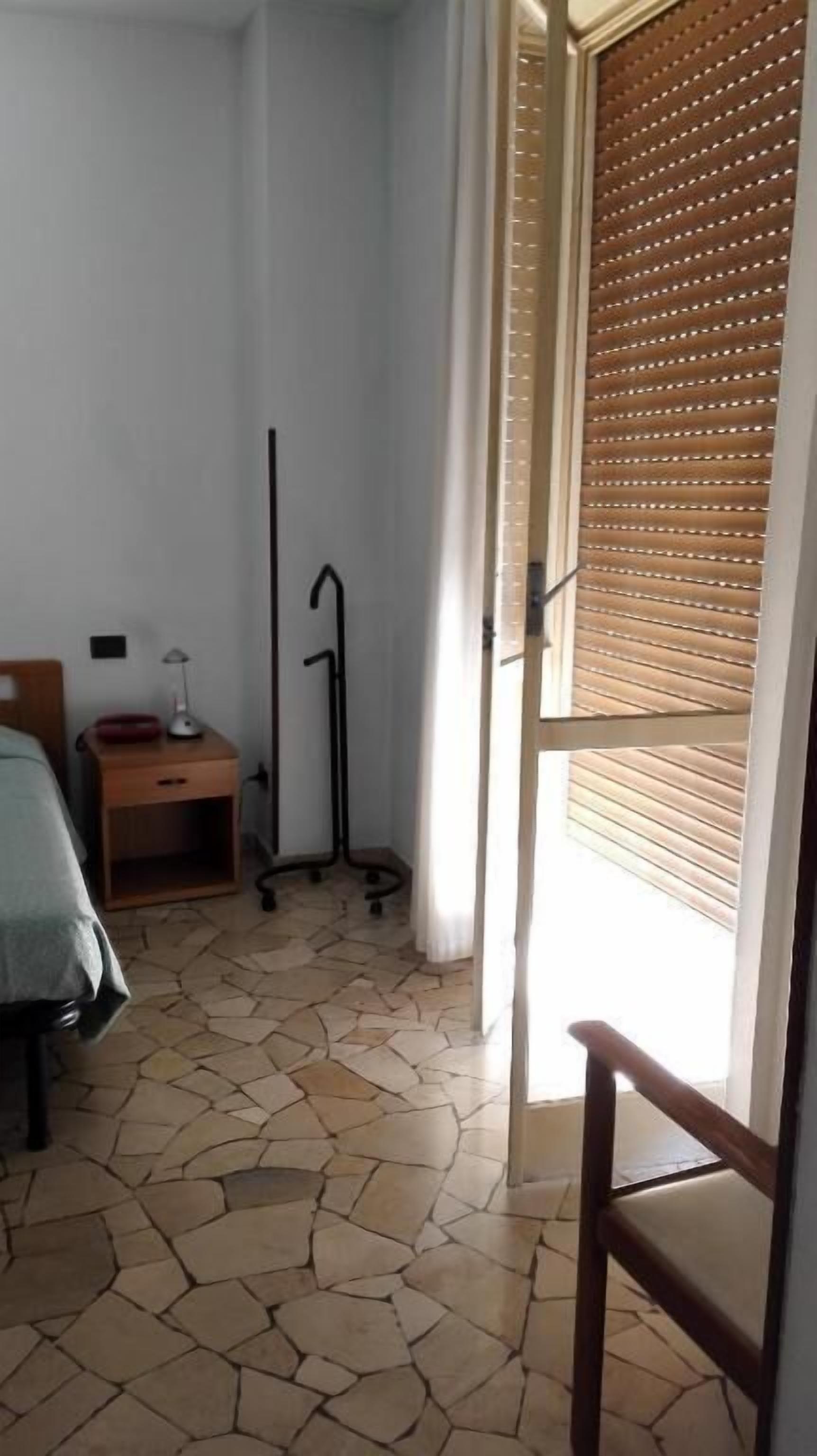 Une chambre d’hôtel avec un lit, une table de chevet avec une lampe, un plancher en bois et une vue par une fenêtre équipée de stores.