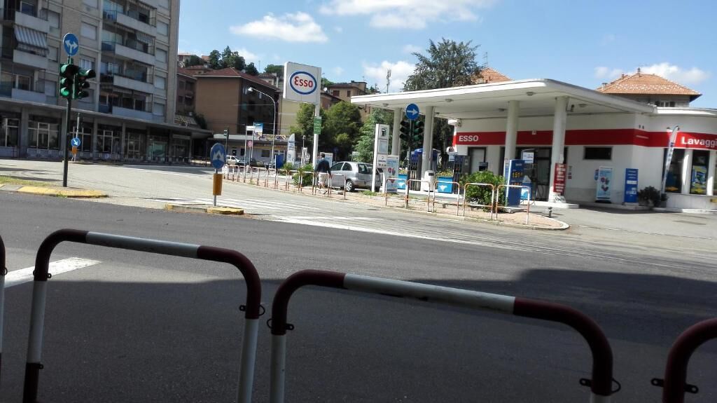 Une station-service avec un logo rouge et blanc, un passage piéton et un immeuble à plusieurs étages en arrière-plan.