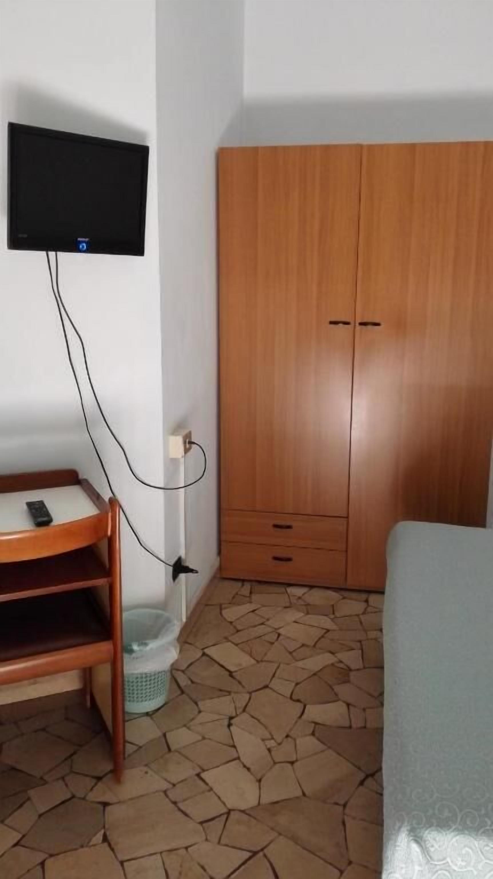 A room with a wooden wardrobe, a television mounted on the wall, a desk with a chair, and a bed.