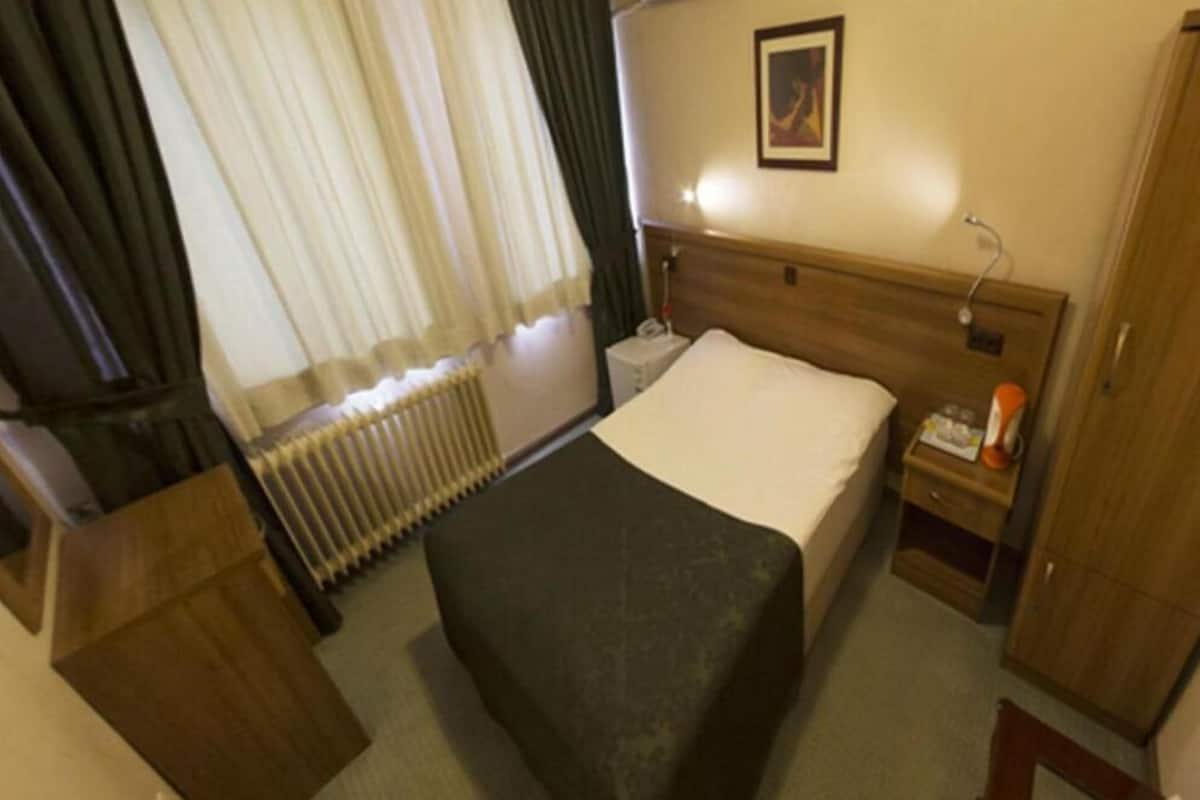 A hotel room with a bed, a bedside table, a nightstand, a radiator, a window with curtains, and a wall-mounted picture.