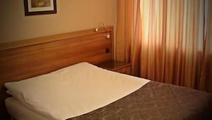 Une chambre d’hôtel avec une tête de lit en bois, un lit simple avec du linge de lit blanc, une couverture marron, un tableau encadré au mur et une fenêtre avec des rideaux.