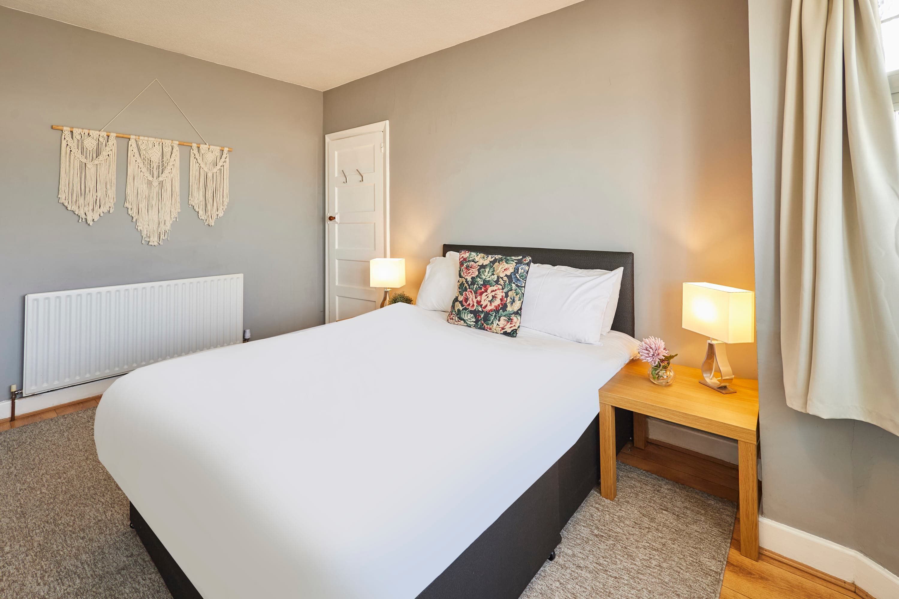 A neatly arranged bedroom with a large bed, bedside lamps, a radiator, and a macrame wall hanging.