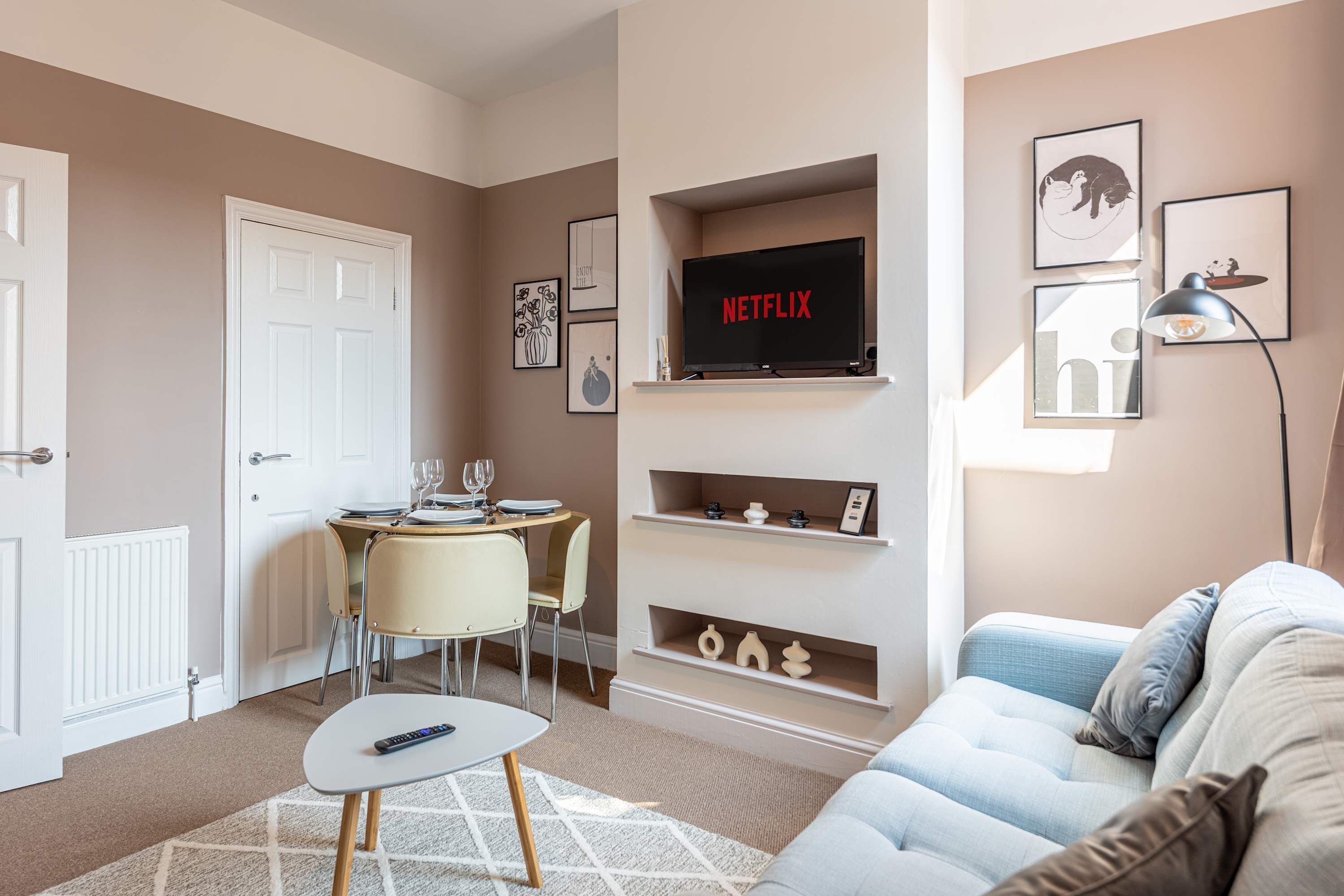 Un soggiorno moderno con un divano, una TV a parete, un'area pranzo con tavolo e sedie e un piccolo scaffale con oggetti decorativi.