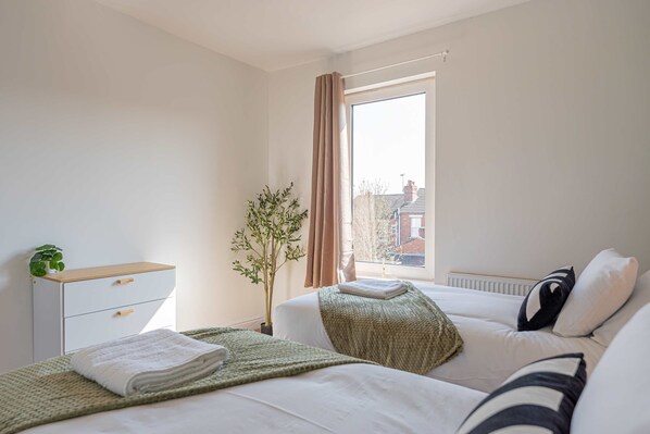 A bedroom with two beds, a window with curtains, a radiator, and a small plant on the dresser.