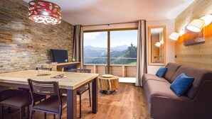 A cozy room with a wooden dining table, chairs, a sofa, and a large window offering a mountain view.