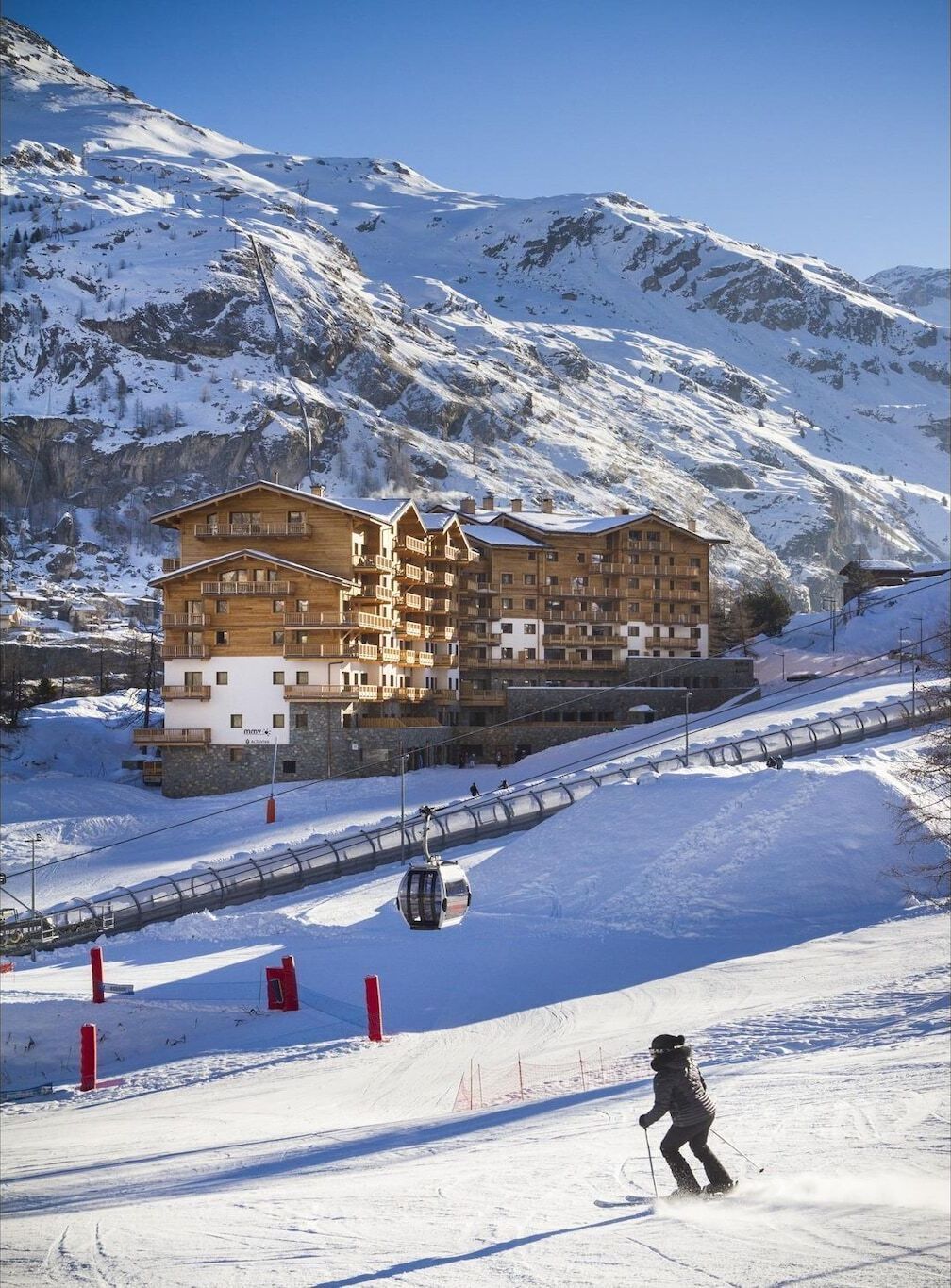 A wooden multi-story building with balconies, likely a hotel, situated on a snowy mountain slope with a ski lift and a skier in the foreground.