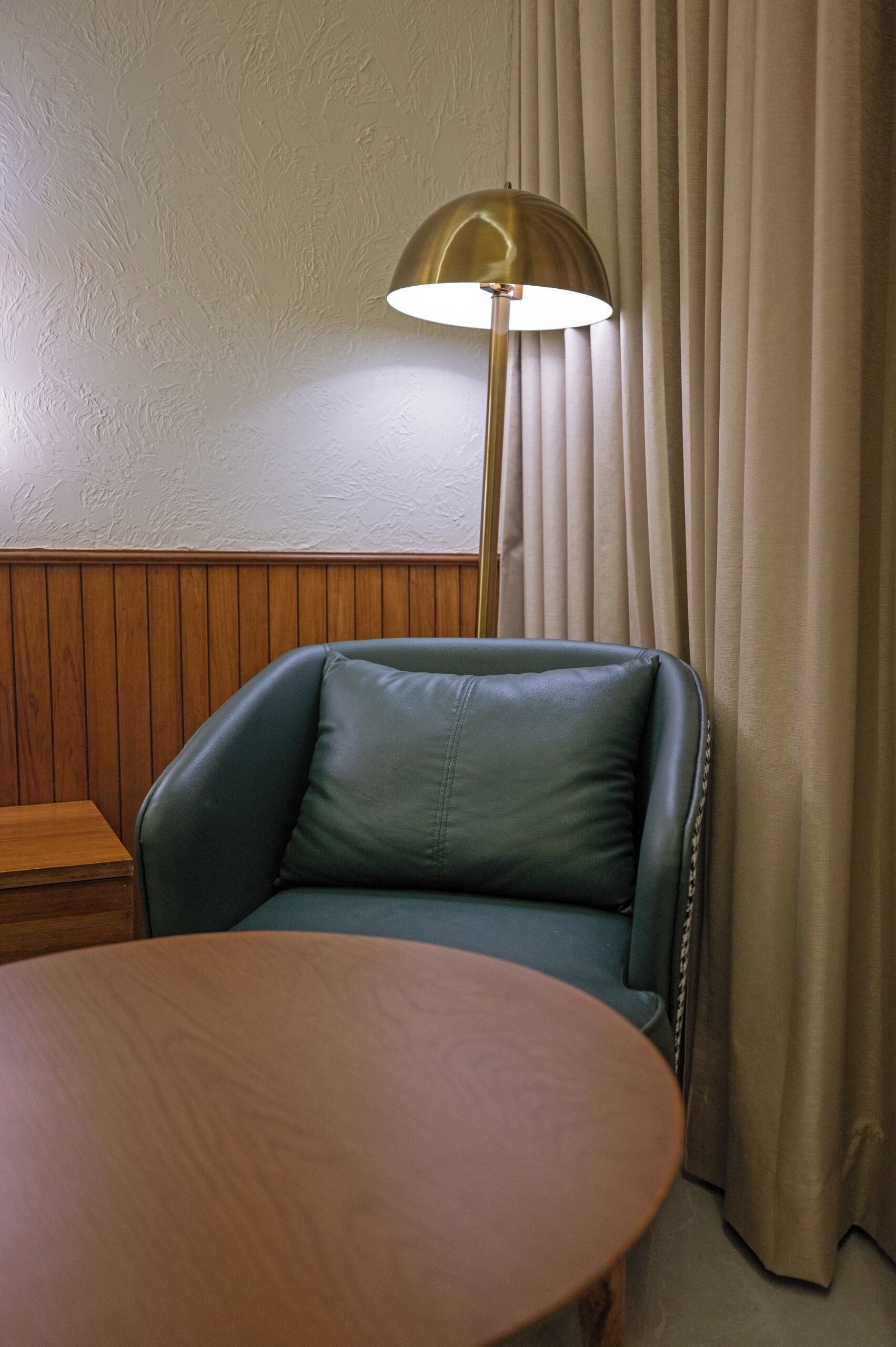 A room with a wooden panel wall, a green leather chair, a round wooden table, and a floor lamp.