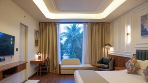 A hotel room with a bed, a desk, a chair, a TV, and a window with a view of palm trees. - Hotel Idyllic Inn (Bhopal)