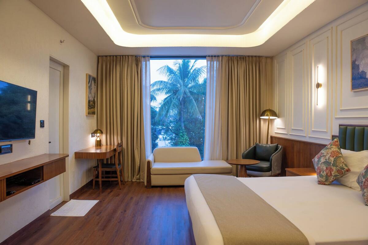 A hotel room with a bed, a desk, a chair, a TV, and a window with a view of palm trees.