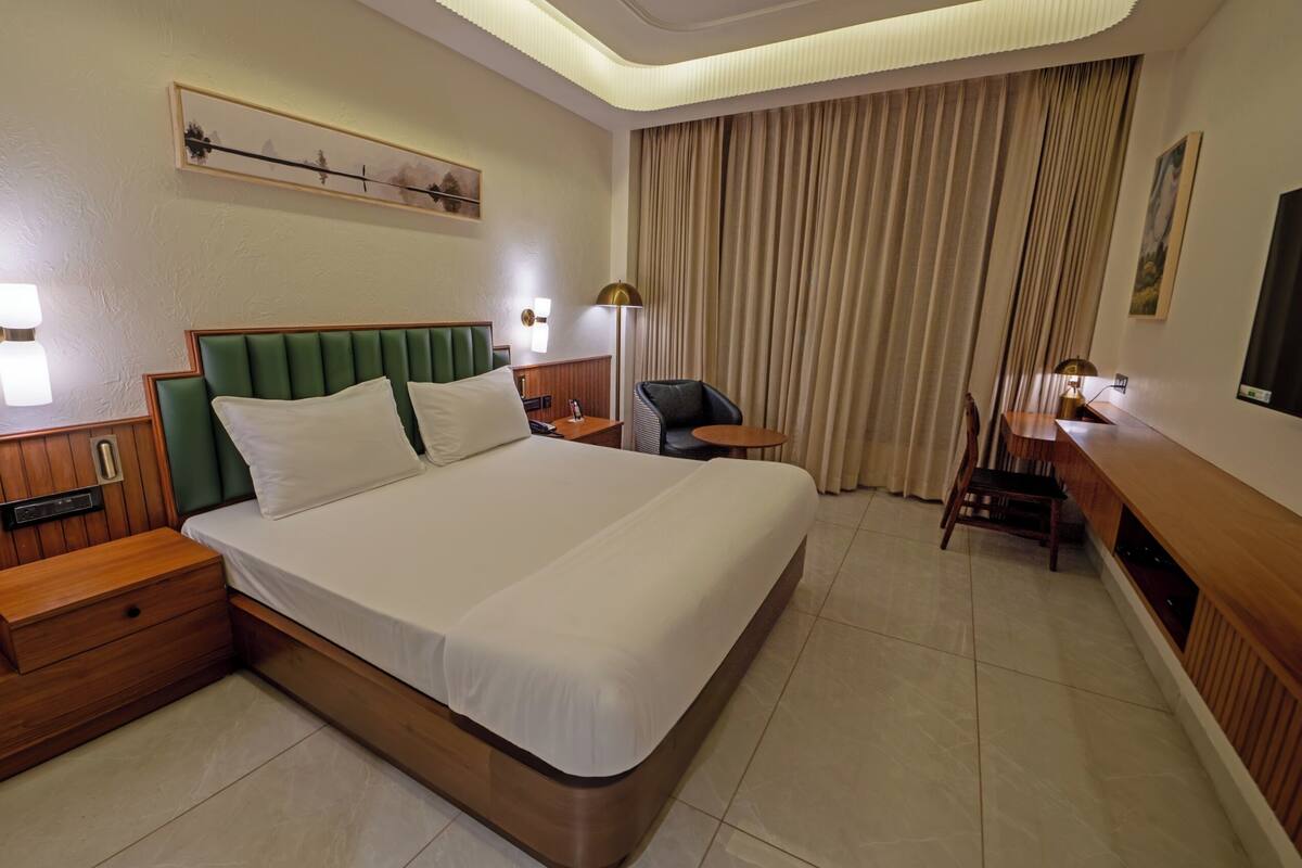 A hotel room with a large bed, wooden nightstands, a desk, a chair, and a large window with curtains.