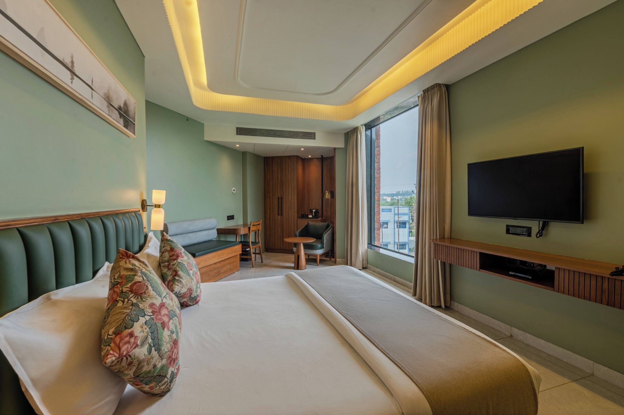 A hotel room with a large bed, a sofa, a small table, a TV, and a window with a view of buildings.