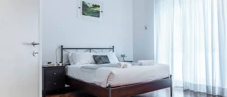 A modern bedroom with a wooden floor, a bed with white bedding, a nightstand, a ceiling fan, and a window with sheer curtains.