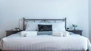 A neatly made bed with white linens, a black leaf-patterned pillow, and two rolled towels. There is a small bedside table with a lamp, a book, and a vase with flowers. A framed picture hangs on the wall above the bed. - Marvellous 2BR flat near city centre (Milan)