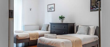 A modern bedroom with two single beds, a dark dresser, a potted plant, and a framed picture on the wall.