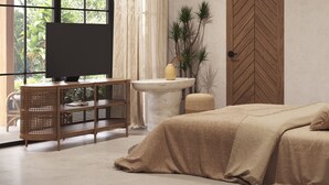 Un dormitorio con un ventanal grande, una puerta de madera, una cama, un televisor sobre un mueble de madera y una mesita con un florero.