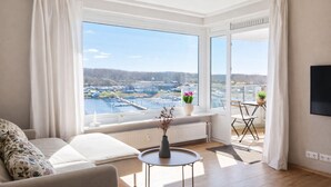 Una sala moderna con un gran ventanal que ofrece vistas a un cuerpo de agua y edificios. Hay un sofá, una mesita con un jarrón y una silla.