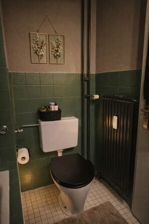 Ein kleines Badezimmer mit Toilette, grünen Fliesen an den Wänden, einem Heizkörper und zwei gerahmten Bildern an der Wand.