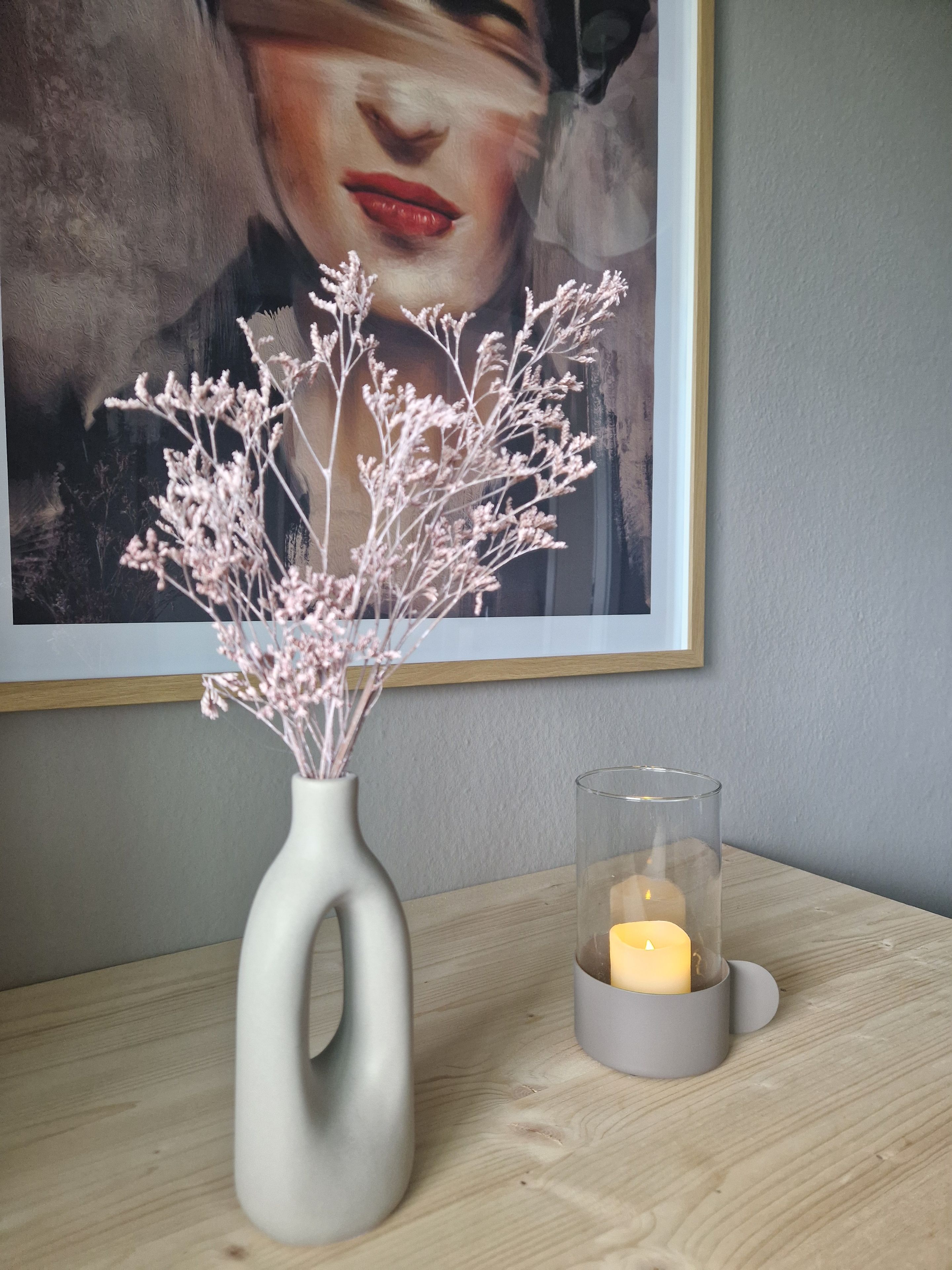 A room with a wooden table, a white vase with dried flowers, and a candle in a glass holder.