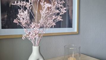 A room with a wooden table, a white vase with dried flowers, and a candle in a glass holder.
