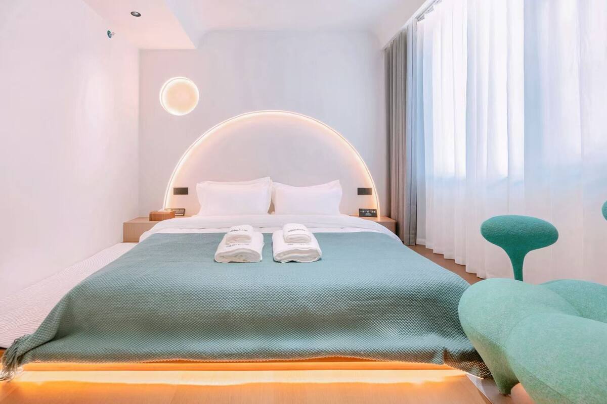 A modern hotel room with a curved headboard, a neatly made bed with two pillows, a green throw blanket, and a teal chair.