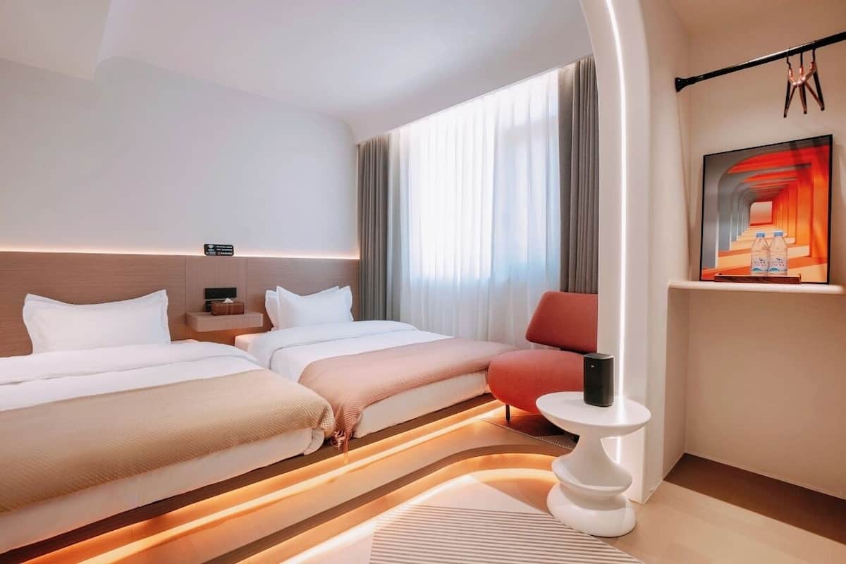 A hotel room with two beds, a small table, a chair, a painting, and a nightstand.