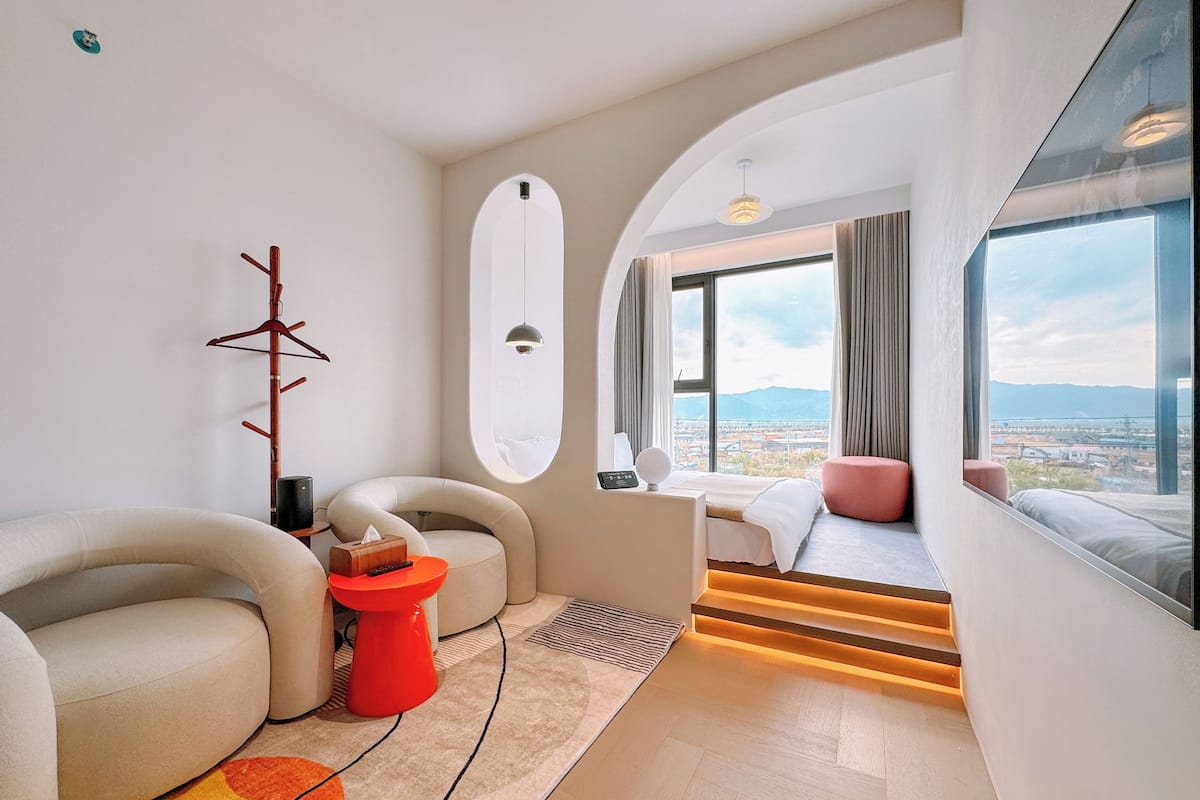 A modern hotel room with a curved sofa, a red stool, a bed with a view, and a large window.