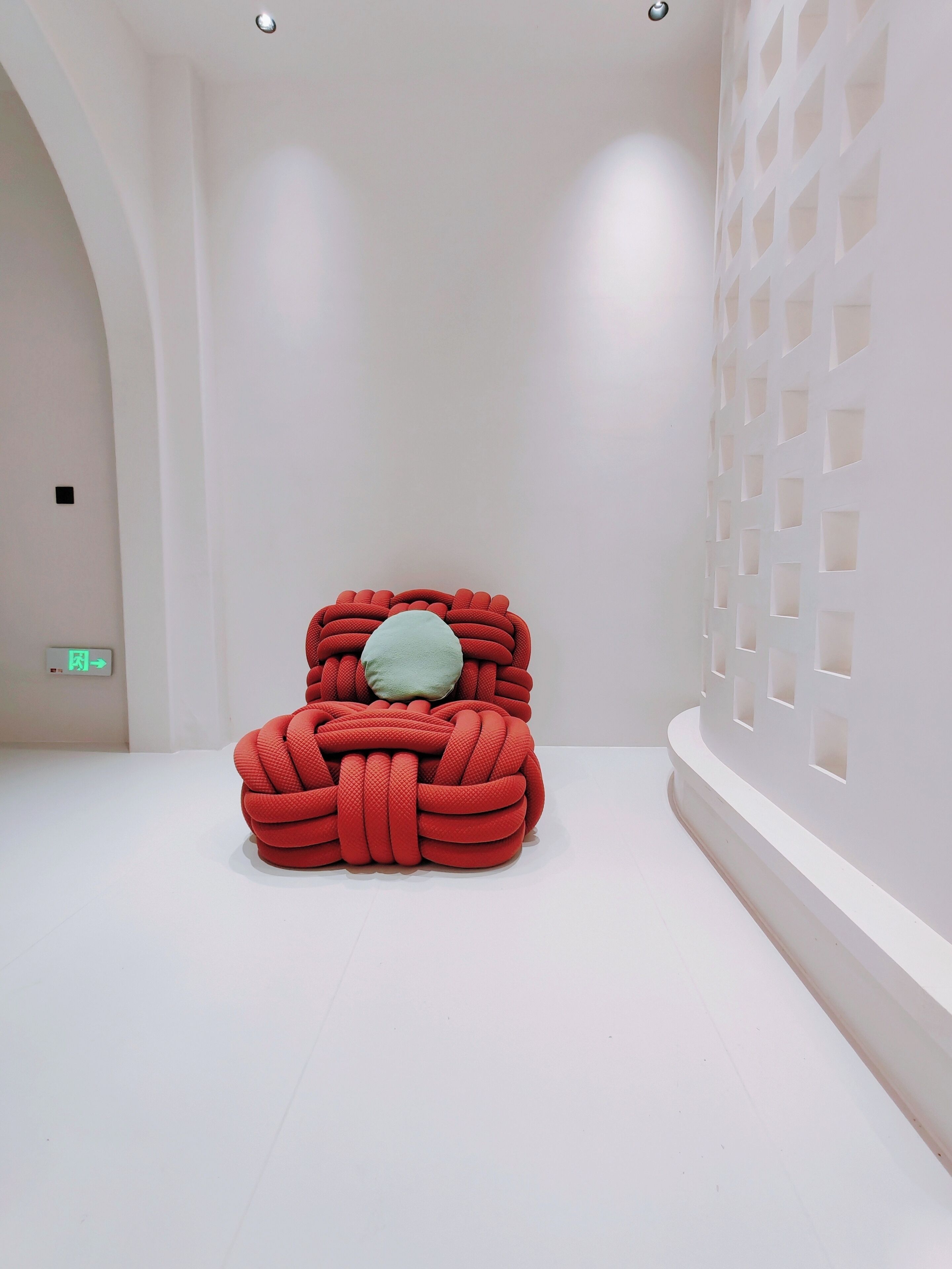 A minimalist room with a unique red, knot-like chair and a light green pillow.