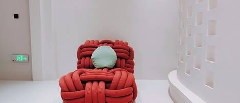 A minimalist room with a unique red, knot-like chair and a light green pillow.