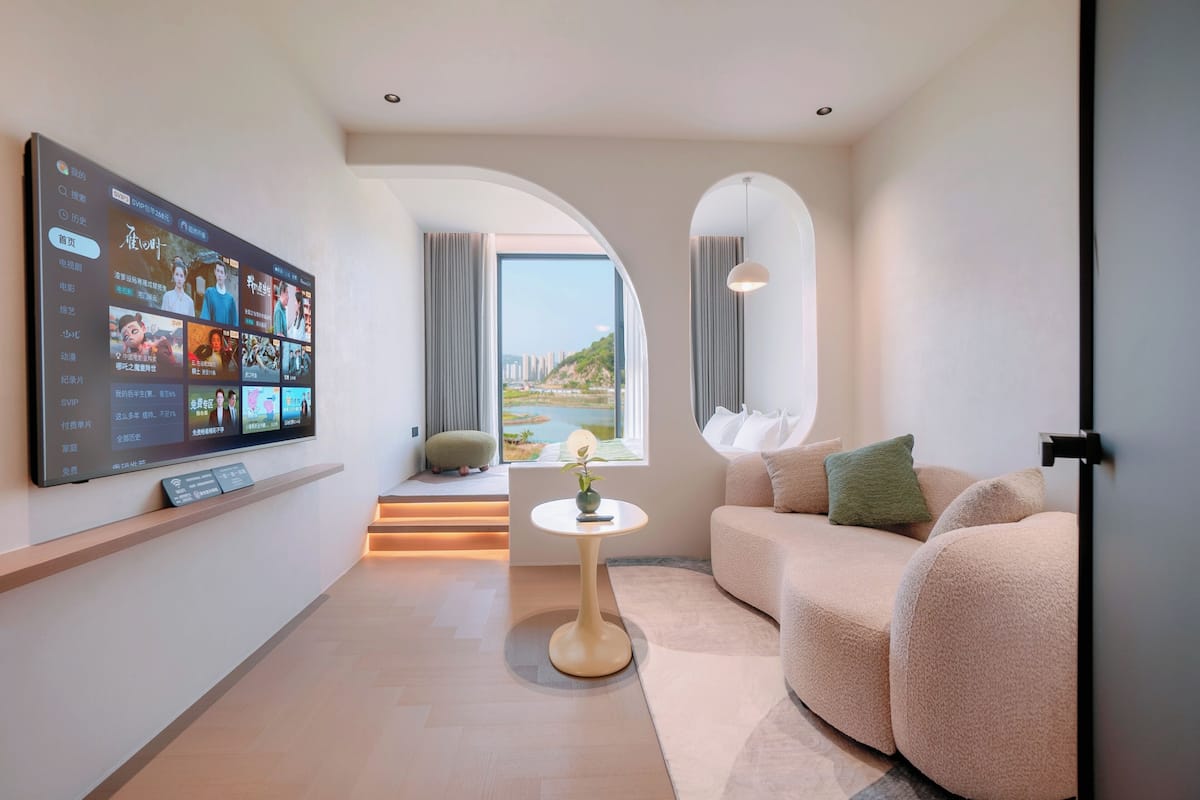 A modern hotel room with a large flat-screen TV, a sofa, a small round table, and a view of a cityscape through an arched window.