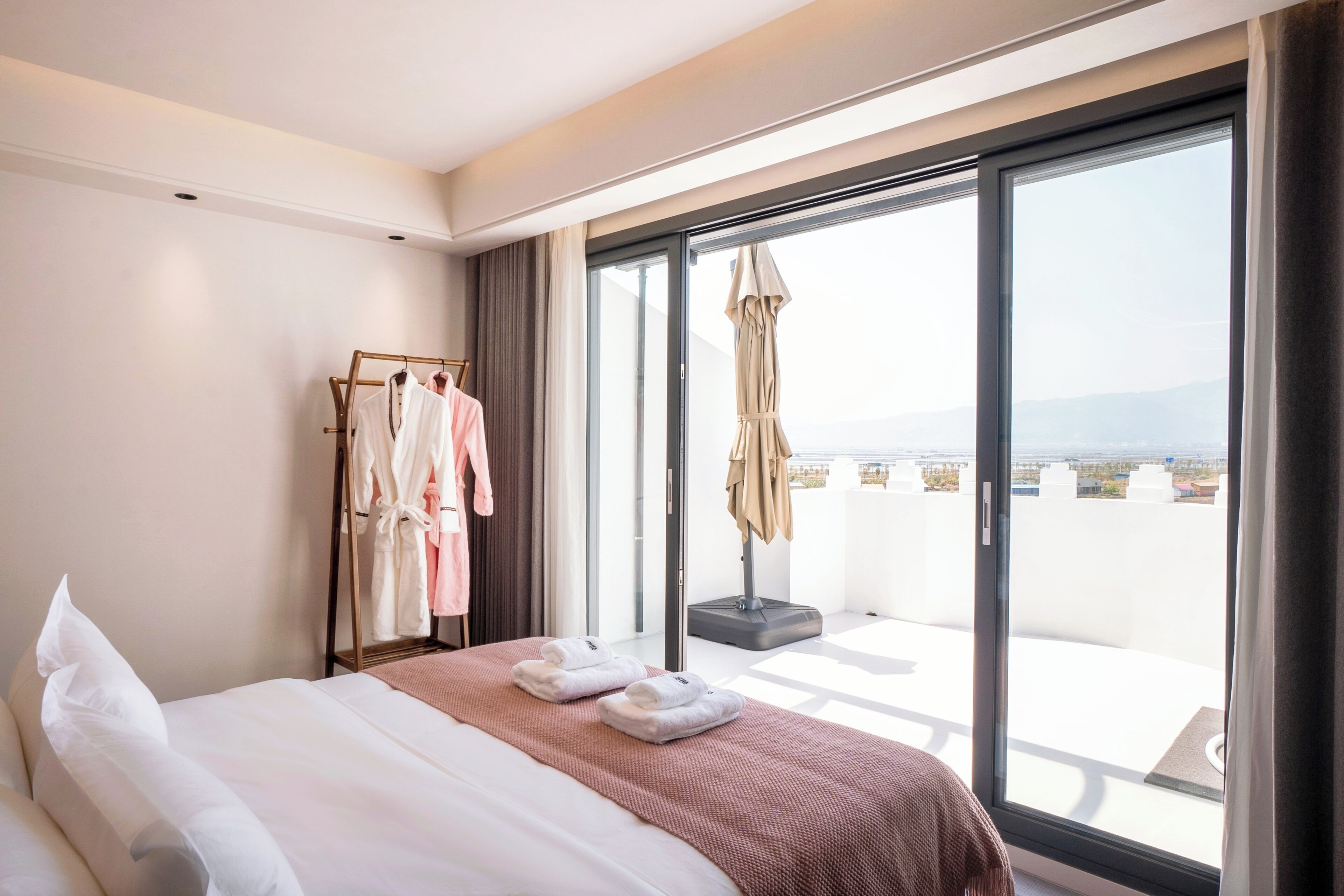 A hotel room with a bed, a robe rack, and a view of the city through large windows.