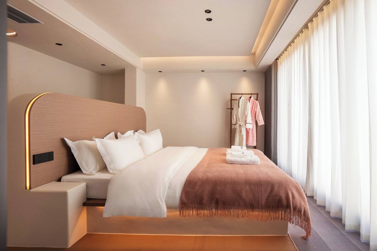 A modern hotel room with a large bed, a bedside table, a wardrobe, and a window with curtains.