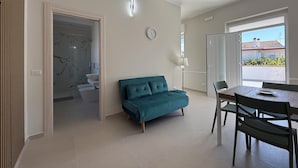 Una sala de estar moderna con un sofá color turquesa, una mesa de comedor con sillas, un ventanal con vistas a un edificio de techo rojo, y un baño con lavamanos y ducha de mármol.