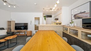 A modern living room with a wooden dining table, a flat-screen TV, and a kitchen area with a brick backsplash and modern appliances.