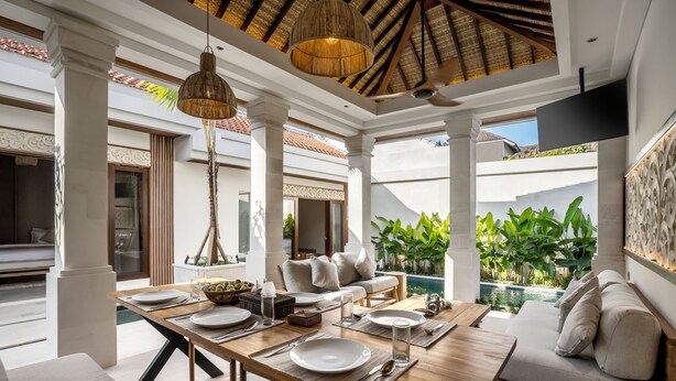 A dining area with a wooden table set for four, surrounded by a white and beige color scheme, with a view of a swimming pool and greenery. - Villa Advaya (Kerobokan)