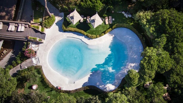 An aerial view of a property with a large circular swimming pool surrounded by lush greenery and a few buildings.