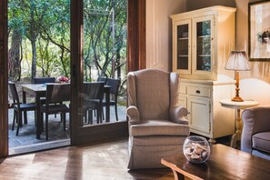 Ein Zimmer mit Blick auf einen Garten, eine Essecke mit Stühlen und Tisch, ein gepolsterter Sessel, ein Schrank und eine Lampe.