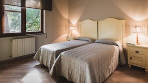 A bedroom with two beds, a window with a view of trees, a radiator, and bedside tables with lamps. - Apartment mit Pool in Strandnähe (Castiglione della Pescaia)