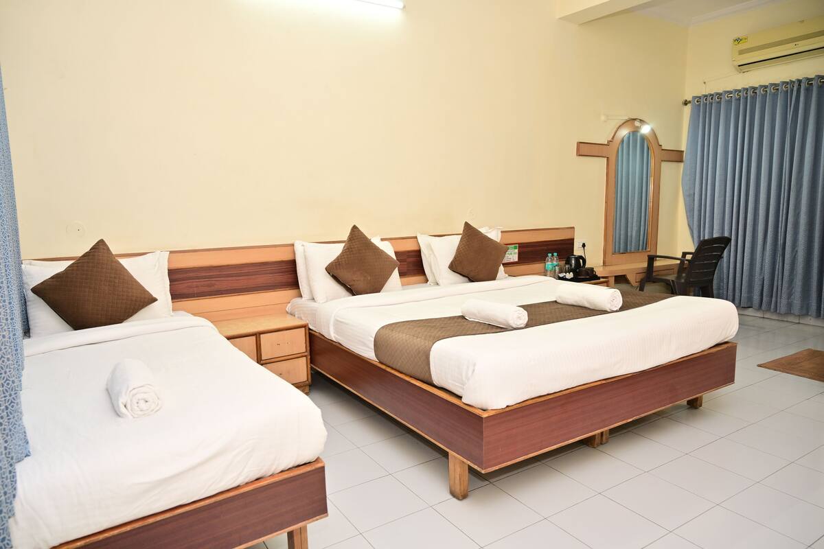 Quarto de hotel com duas camas, cabeceiras de madeira, roupa de cama branca, almofadas castanhas, uma mesa de cabeceira, uma cadeira, uma escrivaninha e um aparelho de ar condicionado.