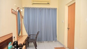 A hotel room with a wooden bench, a small table with a tray of toiletries, a mirror, a window with blue curtains, and a door. - SWAMY RESORT (Shirdi)